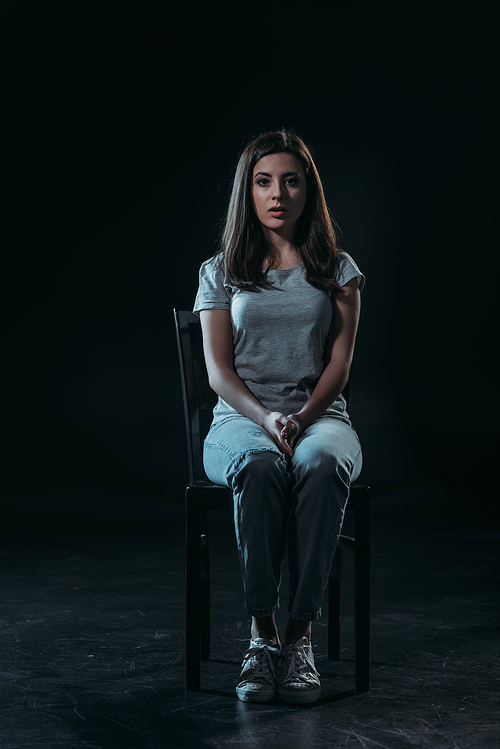 sad young woman  while sitting on chair in darkness on black background