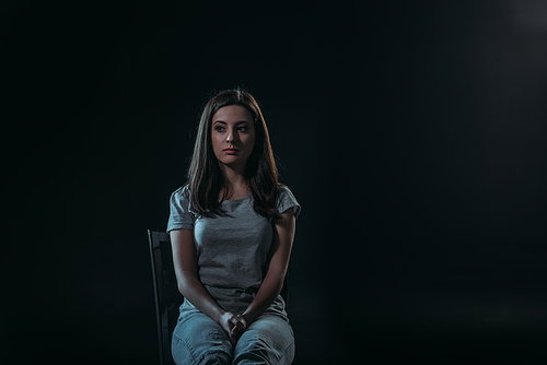 despaired woman looking away while sitting on chair in darkness isolated on black