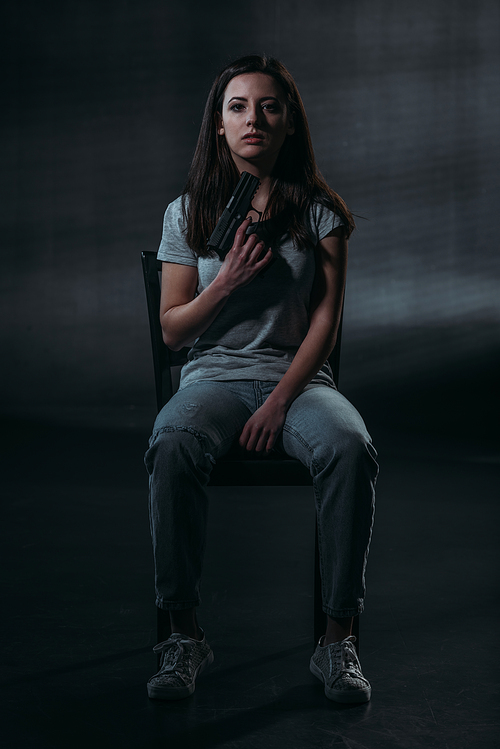 despaired woman holding gun near chin and  while sitting on chair on dark background