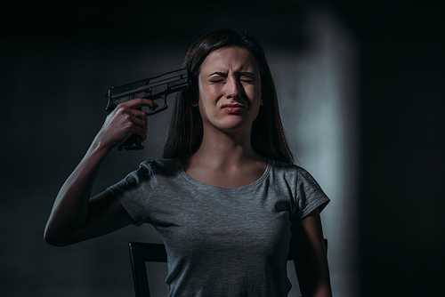 crying, despaired woman holding gun near chin on dark background