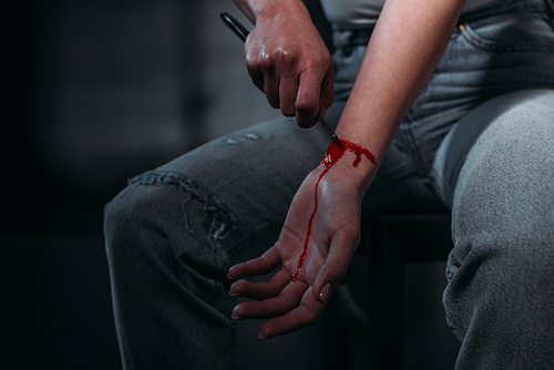 partial view of woman committing suicide by cutting veins with straight razor on dark background