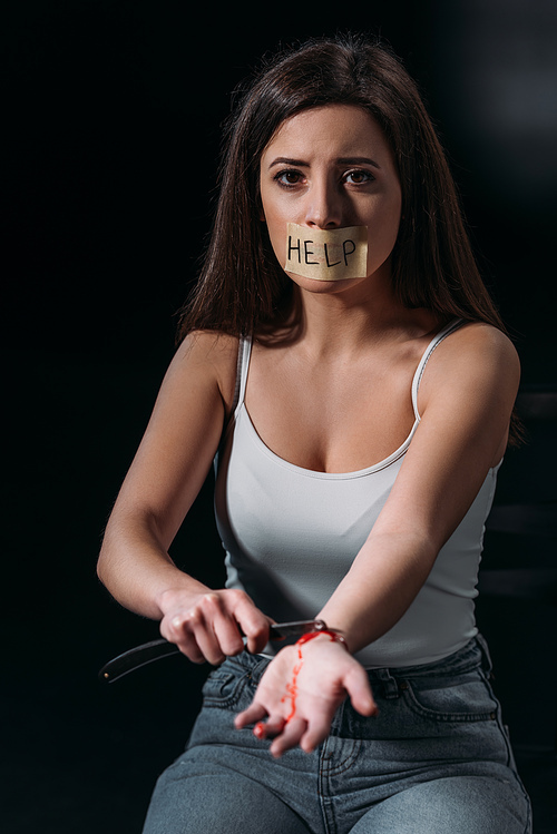 girl with word help on adhesive tape fixed on mouth committing suicide by cutting veins with straight razor on dark background