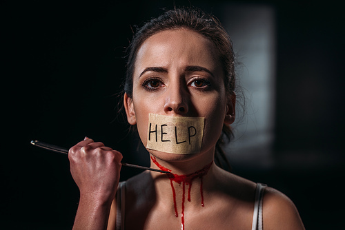 woman with word help on adhesive tape fixed on mouth committing suicide by cutting throat with straight razor on dark background