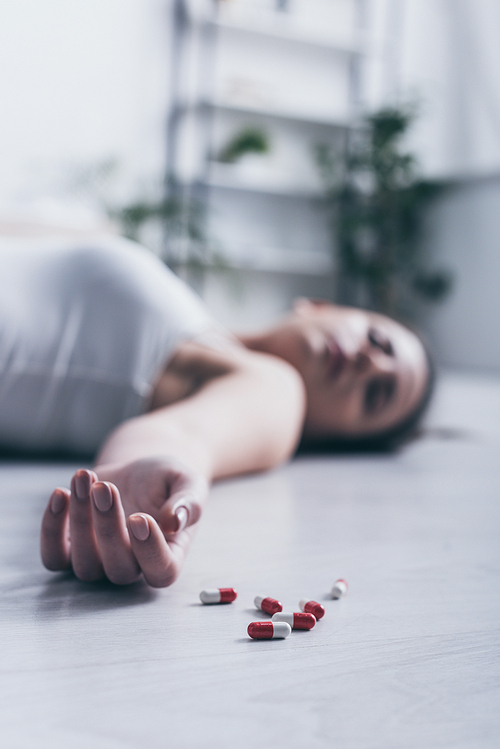 selective focus of lifeless girl, committed suicide by overdosing medicines, lying on floor near scattered pills