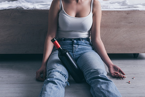 partial view of woman sitting on floor with bottle of alcohol and handful of pills