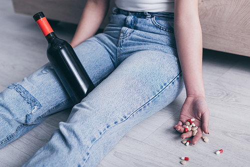 cropped view of unconscious woman sitting on floor with bottle of alcohol and handful of pills