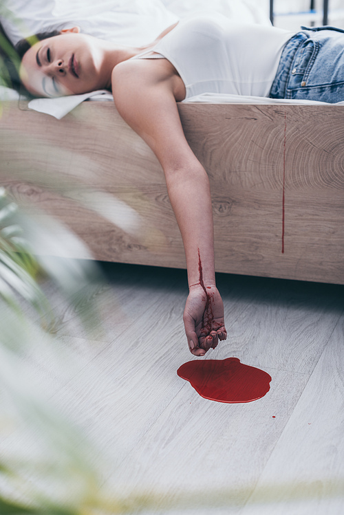 selective focus of lifeless woman with cut bleeding veins lying on bed near puddle of blood on floor