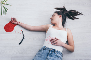 overhead view of lifeless woman with cut bleeding veins lying on floor near straight razor and puddle of blood