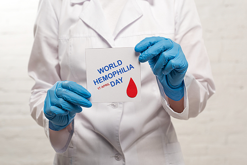 Cropped view of doctor in latex gloves holding card with world hemophilia day on white background