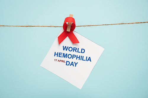 Card with world hemophilia day lettering and red ribbon on cord with pin isolated on blue