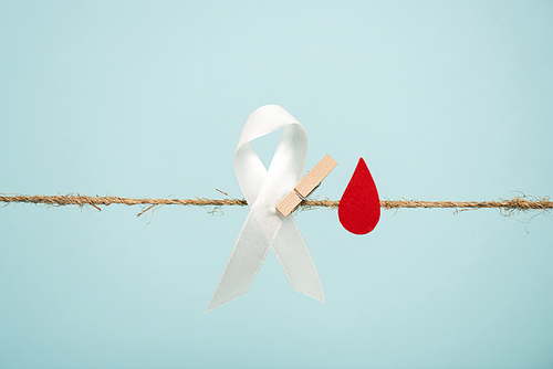 White ribbon with pin and paper in blood drop shape on rope isolated on blue, hemophilia concept