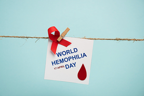 Card with world hemophilia day lettering and red ribbon on rope with pin isolated on blue