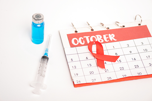 Selective focus of calendar with october month, aids awareness red ribbon with syringe and vaccine on white surface