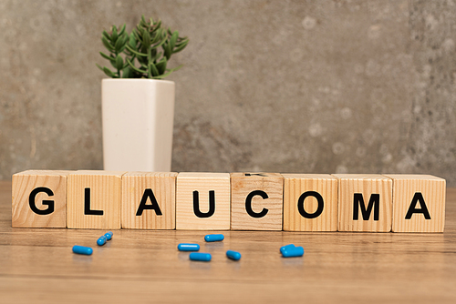 Selective focus of glaucoma lettering on cubes with pills and plant on wooden table on grey background