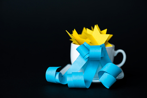 white cup with paper stars near blue ribbons isolated on black