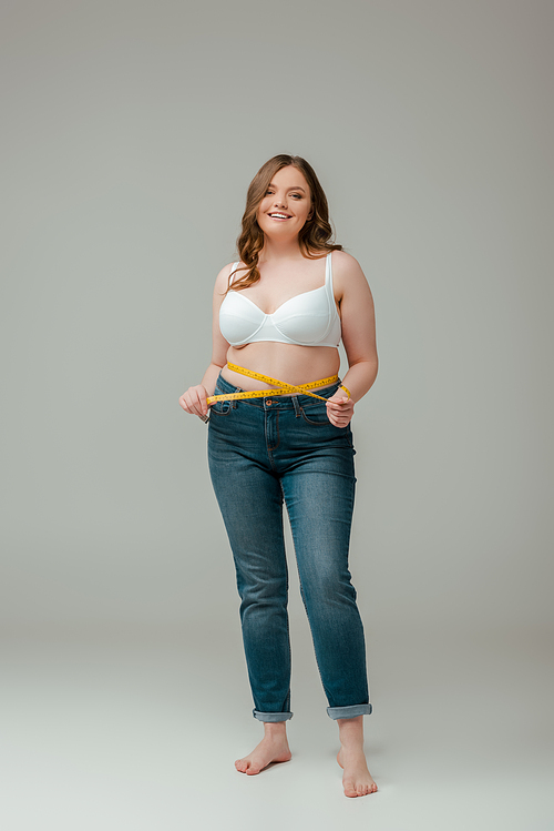 happy plus size woman in jeans and bra measuring waist on grey