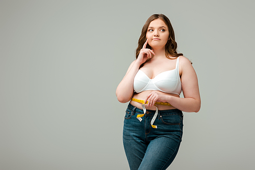 pensive plus size woman in jeans measuring waist isolated on grey