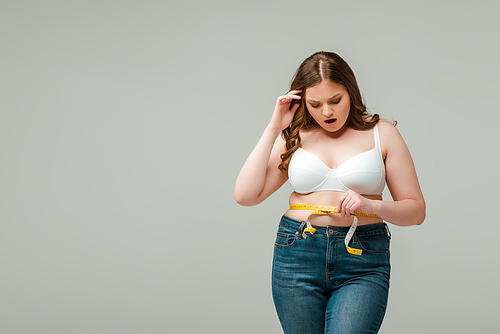 shocked plus size woman in jeans measuring waist isolated on grey