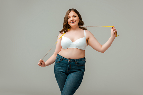 happy plus size woman in jeans holding measuring tape isolated on grey