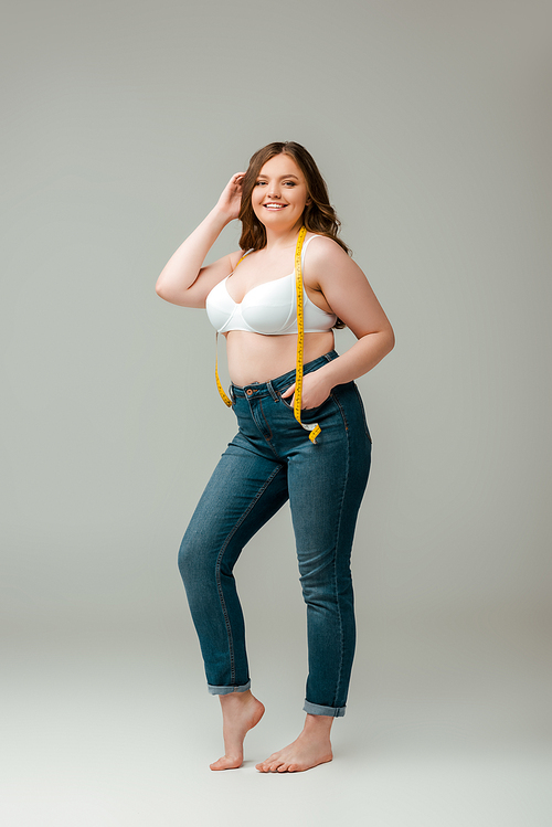 cheerful plus size woman in jeans and bra standing with measuring tape on grey