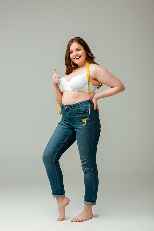 cheerful plus size woman in jeans and bra standing with measuring tape and showing thumb up on grey