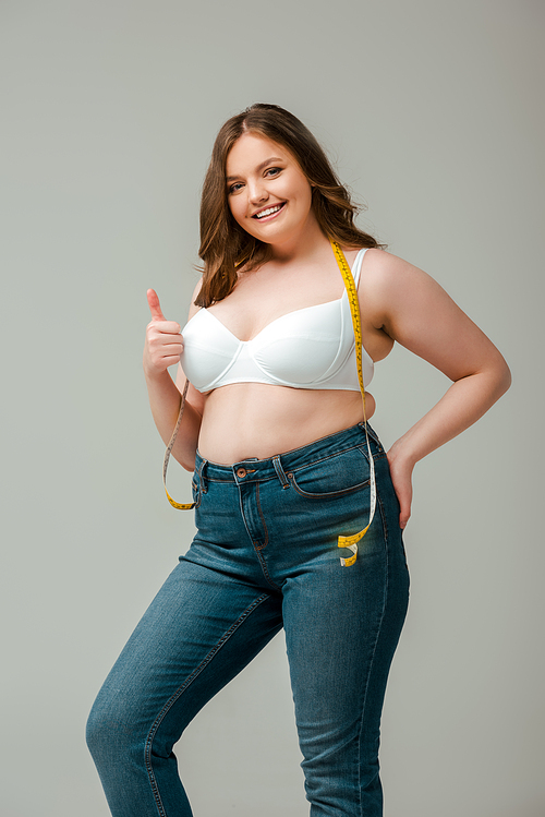 cheerful plus size woman in jeans and bra standing with measuring tape and showing thumb up isolated on grey