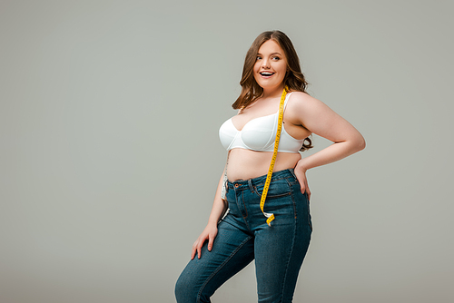 cheerful plus size woman in jeans and bra standing with hand on hip isolated on grey