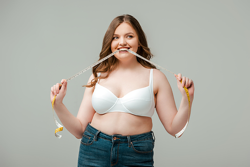 cheerful plus size woman in jeans and bra biting measuring tape and looking away isolated on grey