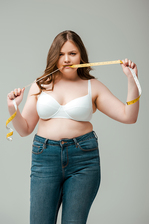 plus size woman in jeans and bra biting measuring tape isolated on grey