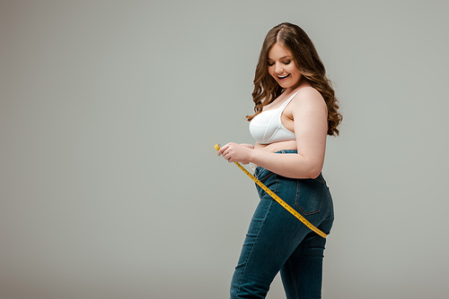 cheerful plus size woman in jeans and bra measuring body isolated on grey