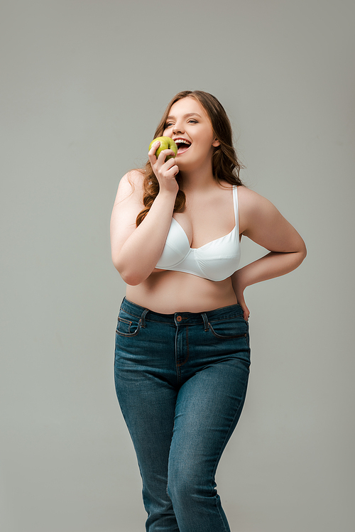 cheerful plus size woman in jeans and bra eating apple isolated on grey