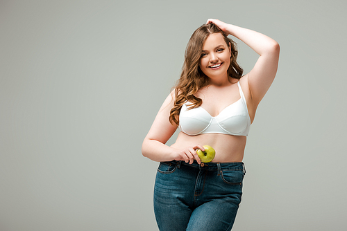 happy plus size girl in jeans and bra holding apple isolated on grey