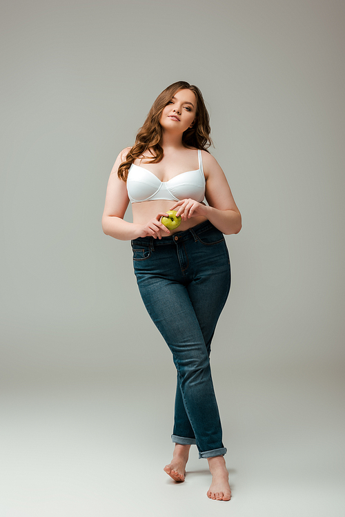 attractive plus size girl in jeans and bra holding apple on grey