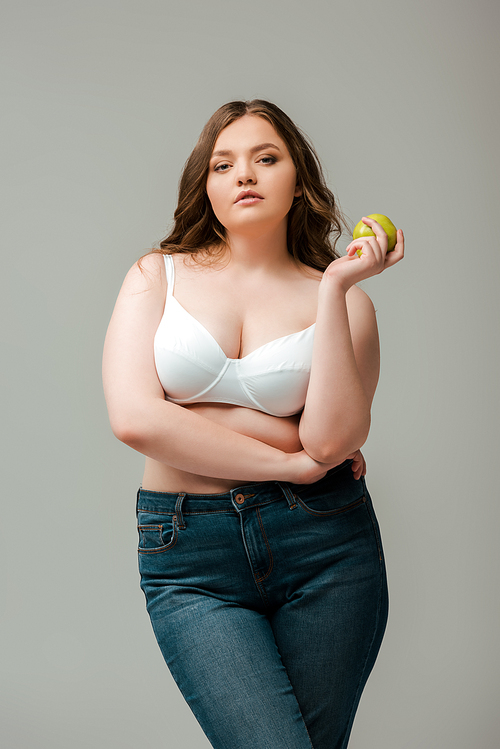 attractive plus size girl in jeans and bra holding apple isolated on grey