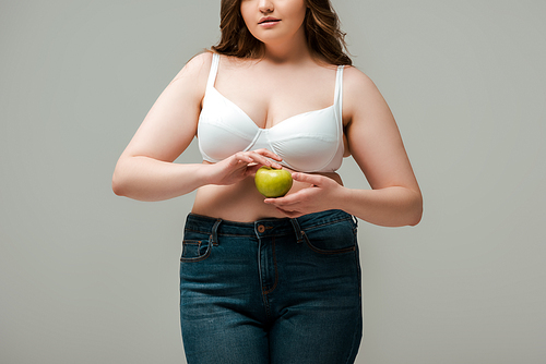 cropped view of plus size girl in jeans and bra holding apple isolated on grey