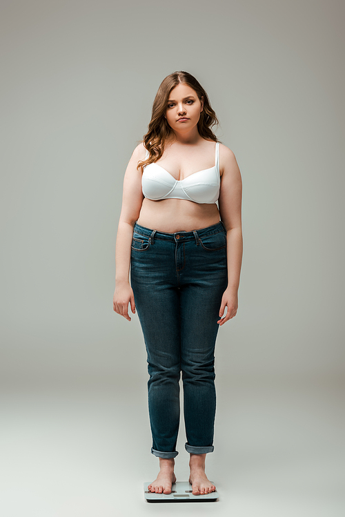 plus size girl in jeans and bra standing on scales and  on grey