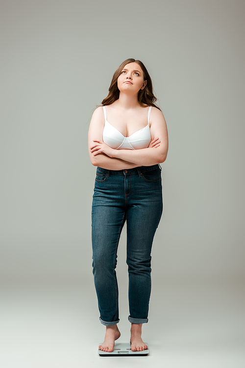 plus size woman in jeans and bra standing with crossed arms on scales and looking up on grey