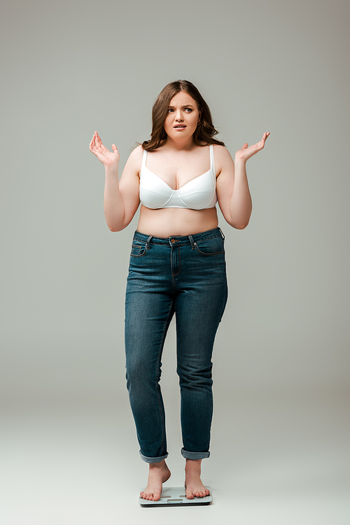 confused plus size girl in jeans and bra standing on scales and gesturing on grey