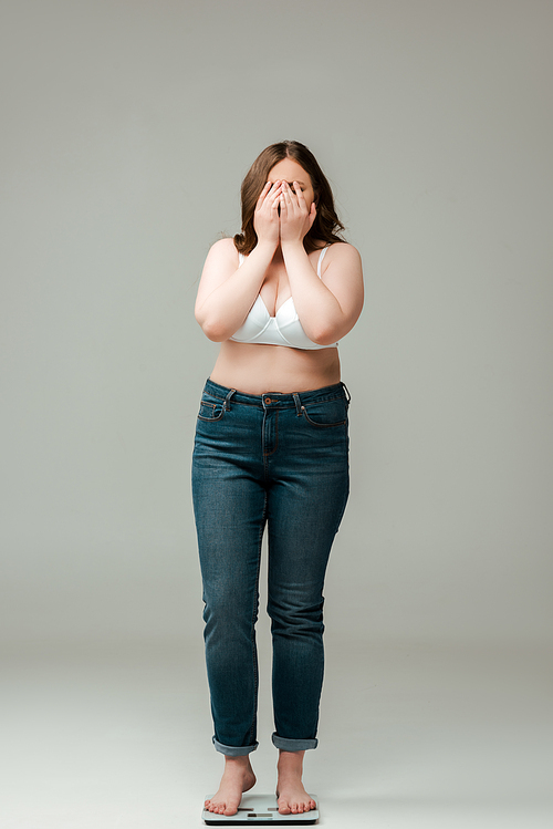 plus size woman in jeans and bra standing on scales and covering face on grey