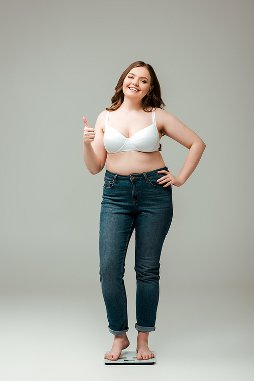 happy plus size woman in jeans and bra standing on scales and showing thumb up on grey