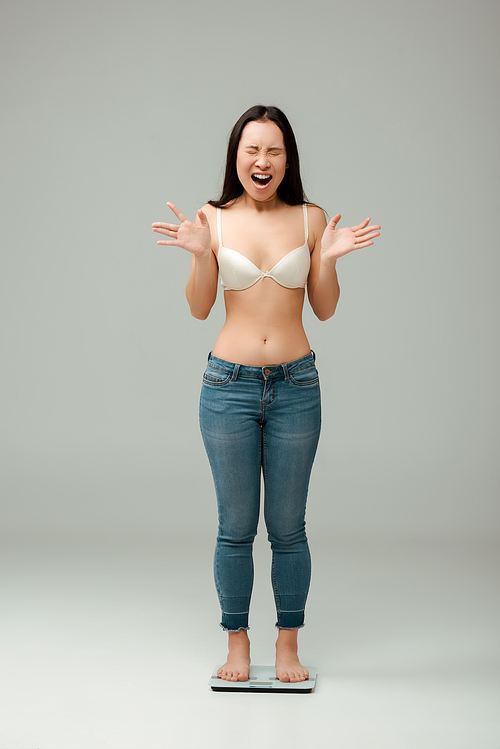 upset asian overweight girl in jeans and bra standing on scales and screaming on grey