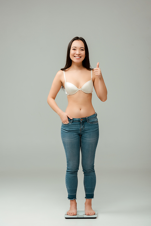 happy overweight asian woman standing on scales and showing thumb up on grey