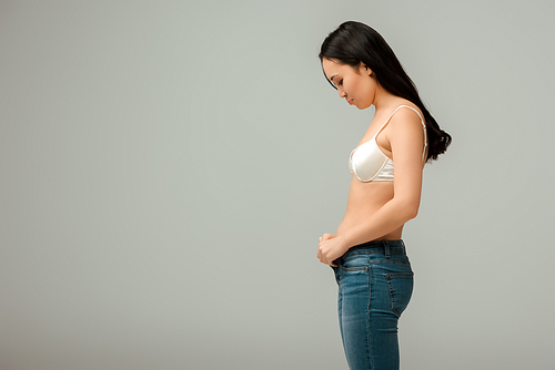 side view of displeased and overweight asian girl wearing jeans isolated on grey