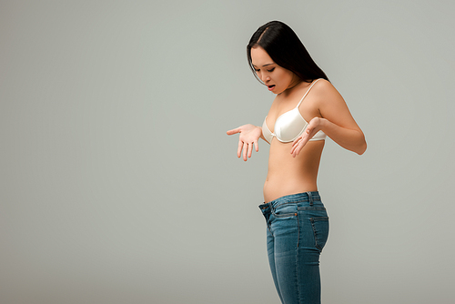 displeased and overweight asian woman gesturing and looking at jeans isolated on grey