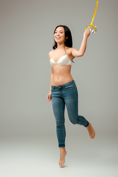 cheerful and overweight asian girl holding measuring tape and jumping on grey