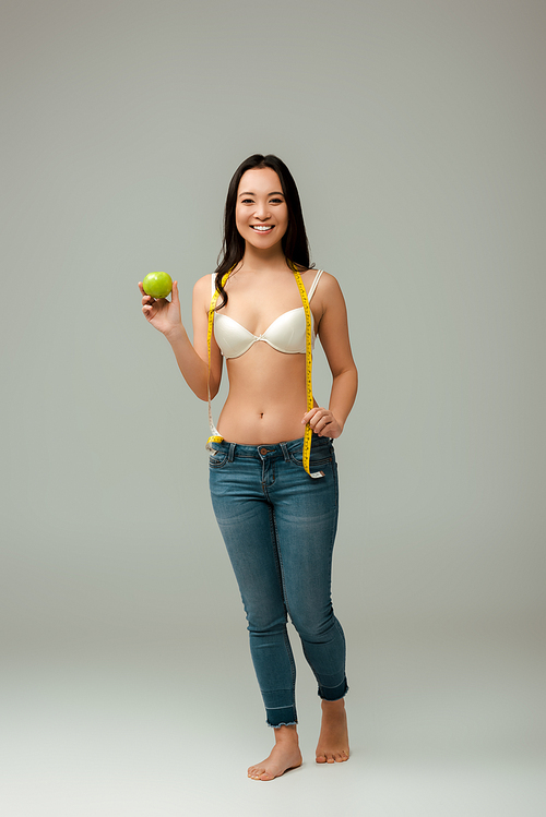 happy and overweight asian girl holding measuring tape and apple on grey