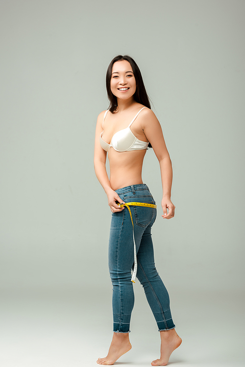 happy and overweight asian girl in bra measuring hips on grey