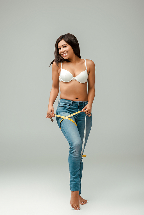 smiling and overweight african american girl in jeans and bra measuring thigh on grey