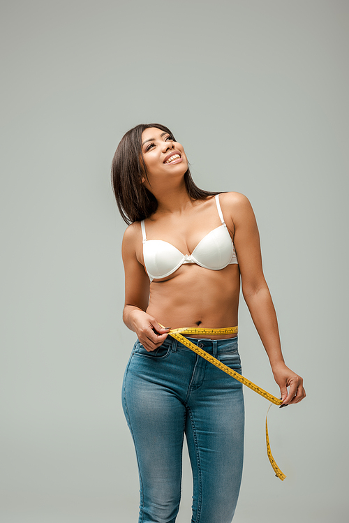 happy and overweight african american girl in jeans and bra measuring waist isolated on grey