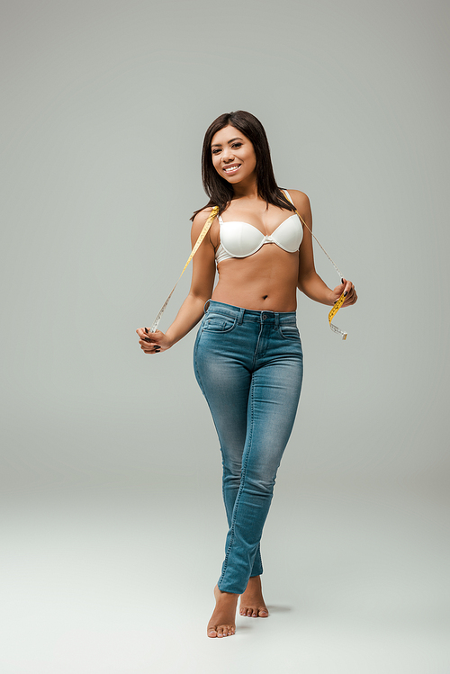 happy and overweight african american girl in jeans and bra holding measuring tape on grey